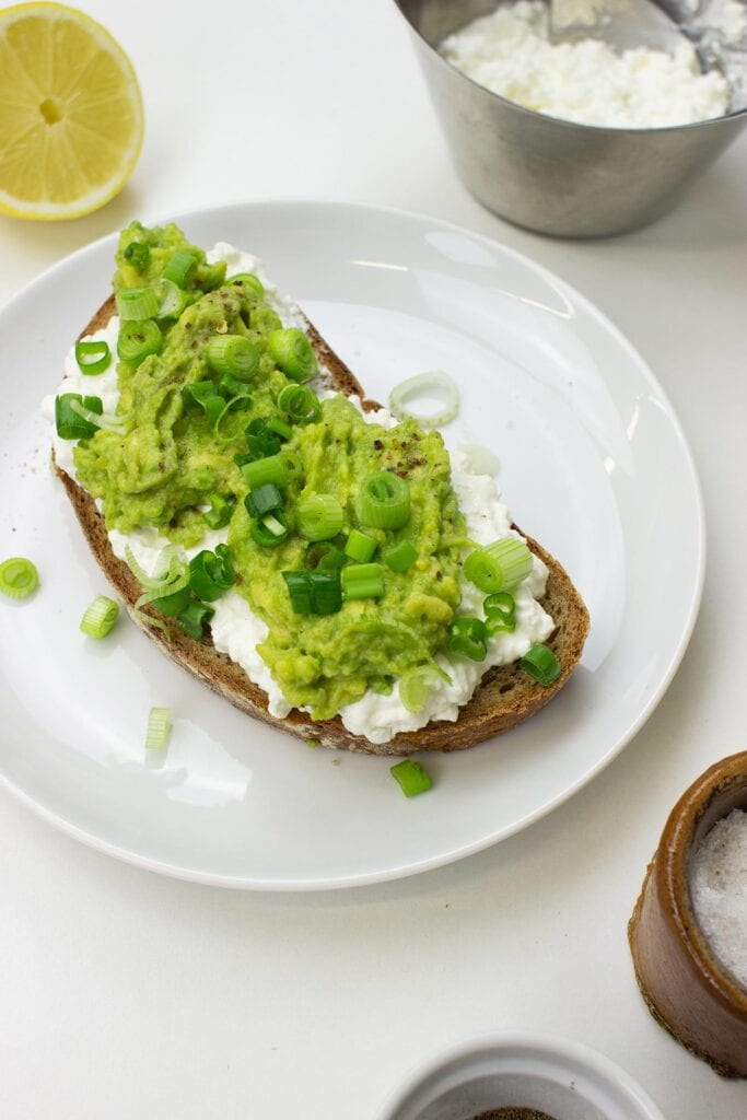 The Avocado Toast with Cottage Cheese is on the plate on a table with salt, pepper, lemon and cottage cheese in the bowl #lemon #green onion | hurrythefoodup.com