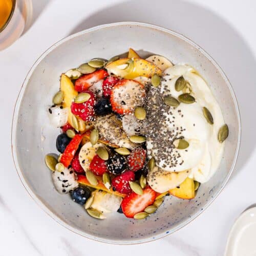 A bowl of fruit salad with yogurt, with a spoon, a glass of tea and some pumpkin seeds. | Hurry The Food Up