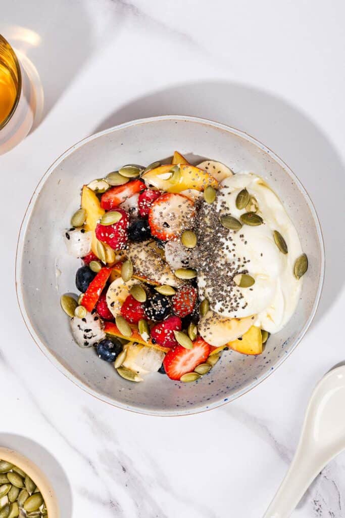 A bowl of fruit salad with yogurt, with a spoon, a glass of tea and some pumpkin seeds. | Hurry The Food Up