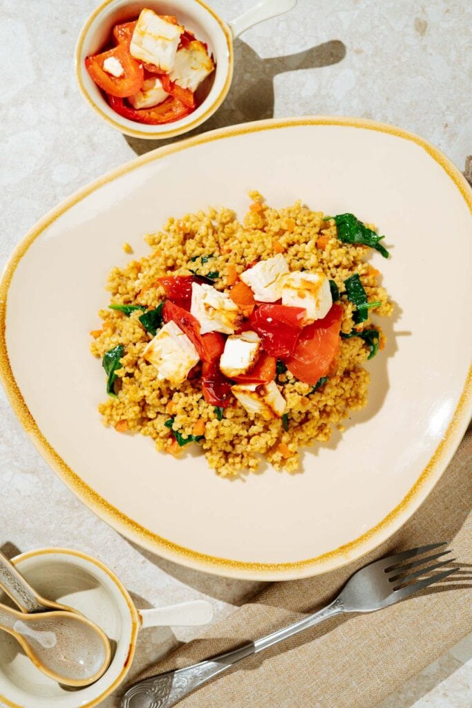 Easy high-protein millet pilaf is on the light plate with baked feta on th top of it. Near it there are the dish with baked vegetablea and feta, two spoons, fork etc | Hurry The Food Up
