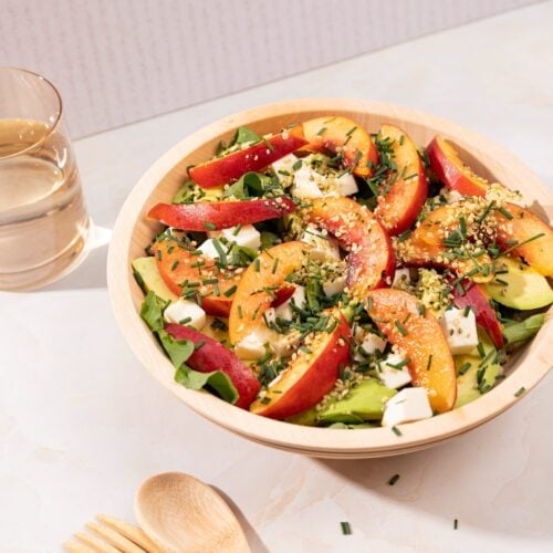 The nectarine salad is in the wooden bowl. There are a glass of water and wooden spoon and fork next to it | Hurry The Food Up