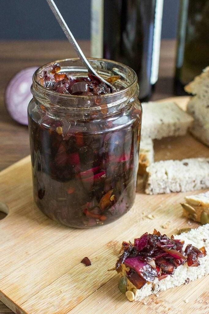 A jar of red onion chutney with a spoon in it, sat on a wooden chopping board. Next to the board are some slices of bread | Hurry The Food Up