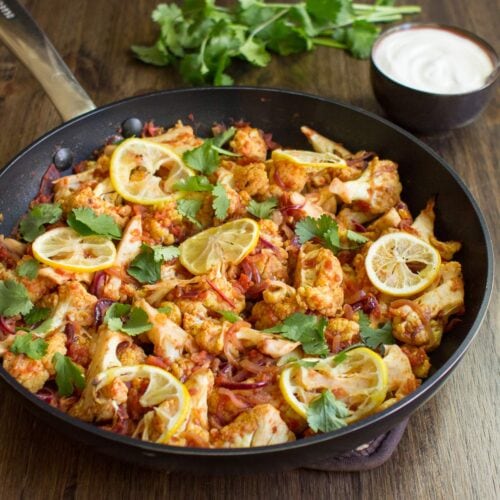 Roasted Cauliflower Curry is in the pan on the wooden surface. Next to it there is a plate with yogurt and fresh sprig cilantro | Hurry The Food Up