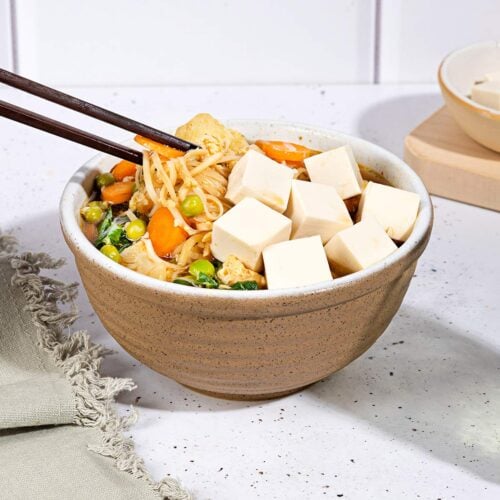 A person is picking up noodles from the bowl of egg drop soup that is on the white table along the bowl with tofu cubes, grey towel and a glass of water | Hurry The Food Up