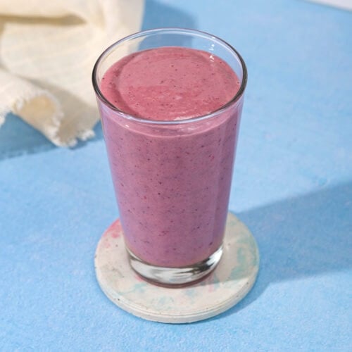 In the centre of a blue table is a white ceramic coaster with a tall glass full of blackberry smoothie. Next to the glass is a linen cloth | Hurry The Food Up