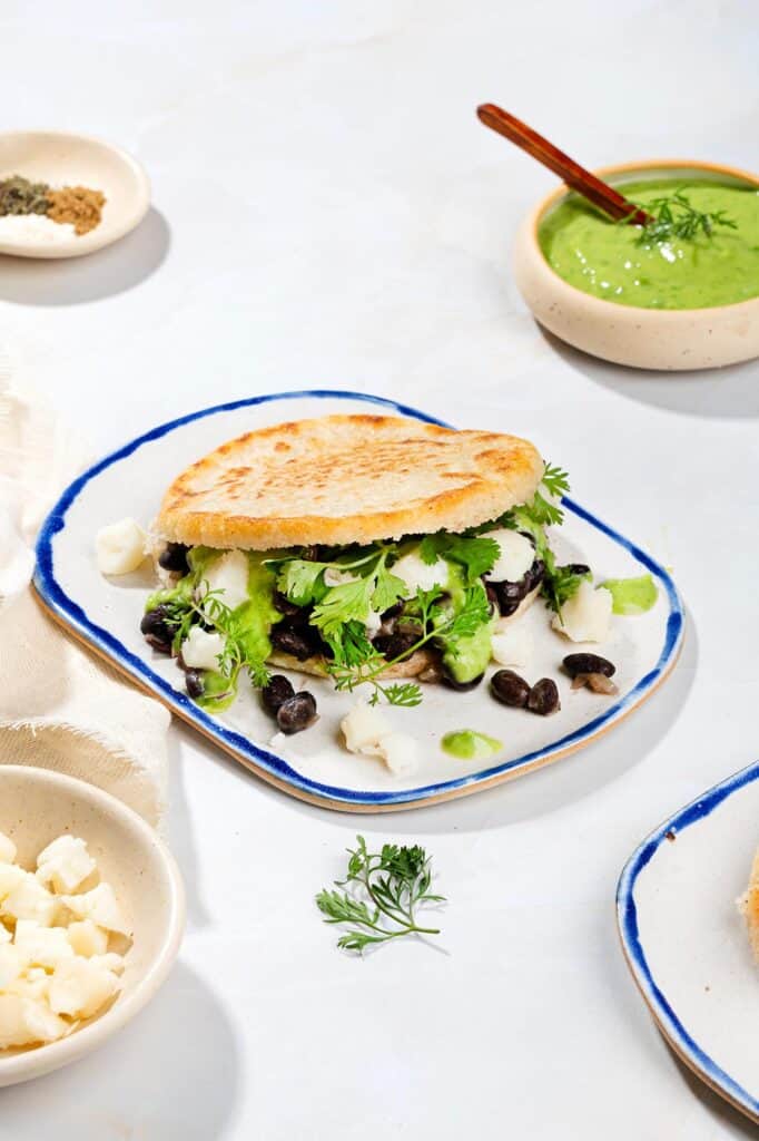 Arepas with black beans on a plate, with a bowl of green sauce, a bowl of crumbled feta and spices | Hurry The Food Up